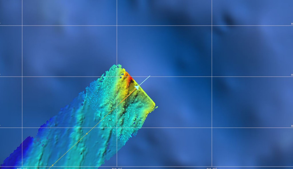 Screenshot from the multi-beam operating system during our transit to the TAG hydrothermal field. The global satellite altimetry dataset is in blue in the background with a spatial resolution of more than a kilometre. In colour is our recorded, ship-based echo-sounder data in a significantly higher resolution (less than 100 m) imaging the seafloor in much more detail. The ship track is in orange and the position and heading of the Meteor is marked by the blue arrow. / Bildschirmfoto aus der Fächerecholot-Software während unseres Transits zum TAG Hydrothermalfeld. Im Hintergrund, in blau, liegt der Satellitenaltimetrie-Datensatz mit einer Auflösung von mehreren Kilometern. Darüber liegen unsere frisch aufgezeichneten, schiffsbasierten Daten mit einer deutlich höheren Auflösung (weniger als 100 m), die den Meeresboden mit viel mehr Details abbildet. Unser gefahrener Kurs ist in orange eingezeichnet und der blaue Pfeil markiert die Position und momentane Ausrichtung der Meteor. (© Meike Klischies)