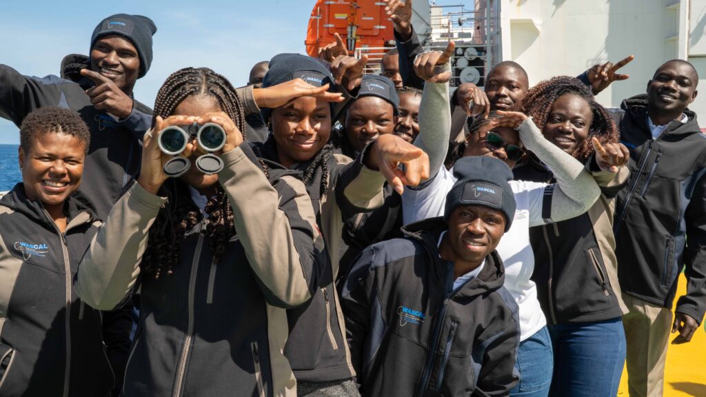 Menschen schauen in die Kamera, eine Person hält ein Fernglas, Schiff im Hintergrund